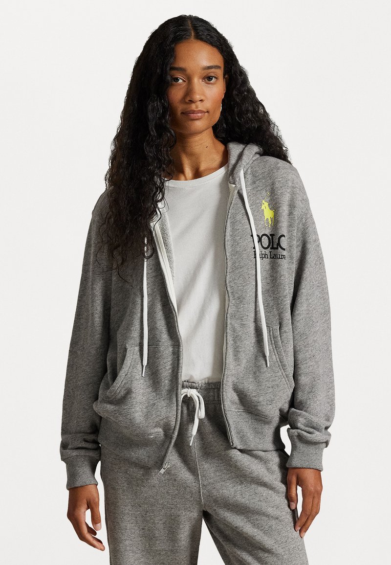 Pelēks zip-up kapuce izgatavota no mīksta auduma, ar baltu auklu, sānu kabatām un dzeltenu logo uz kreisā krūškurvja. Apvienots ar pelēkiem sporta biksēm.