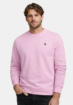 Man with light brown hair wearing a pastel pink sweatshirt with a small black embroidered logo on the chest, hands in pockets.