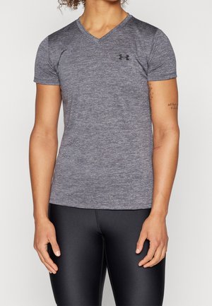 Personne portant un t-shirt Under Armour gris chiné à manches courtes et col en V, et un legging noir moulant, bras reposant le long du corps.