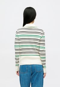 Femeie cu părul lung, drept și închis la culoare, purtând un pulover alb cu dungi orizontale negre și verzi și blugi, întoarsă cu fața la o fundal alb.