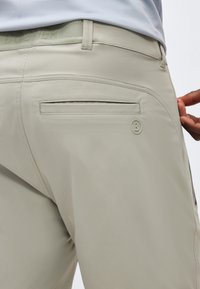 Ανοιχτό μπεζ παντελόνι με πίσω τσέπη με επένδυση, θηλιές για ζώνη και ένα μικρό κυκλικό λογότυπο κοντά στην τσέπη, φορεμένο με ένα πουκάμισο μέσα από το παντελόνι.