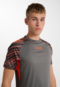 Jonge man met kort blond haar, gekleed in een grijs-oranje sportshirt met patroon en het "hummel"-logo op de borst, kijkt recht vooruit tegen een witte achtergrond.