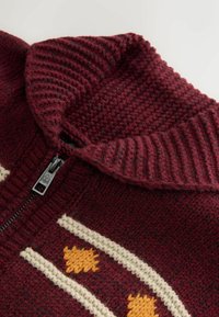 Bordeauxfarbener Strickpullover mit hohem Kragen, der cremefarbene und orangefarbene Muster sowie einen schwarzen Reißverschluss aufweist. Strukturierter Stoff mit gerippten Akzenten.