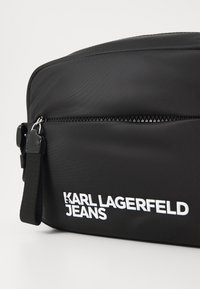 Schwarze Nylon-Umhängetasche mit glatter Textur, ausgestattet mit einem weißen Logo "KARL LAGERFELD JEANS" und einem Reißverschluss. Verstellbarer Schulterriemen inklusive.