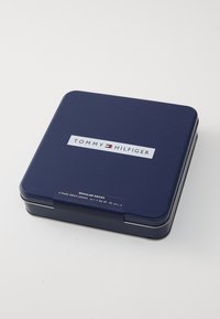Scatola metallica blu navy con una texture liscia, dotata di un'etichetta con il logo "TOMMY HILFIGER" e un tag che indica "4 paia di calzini da uomo."