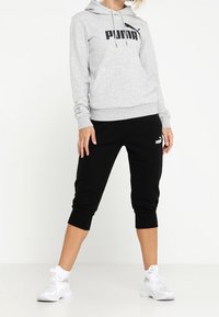 Sweat à capuche gris avec logo noir "PUMA", associé à un pantalon de survêtement noir raccourci avec un petit logo. Chaussures de sport blanches.
