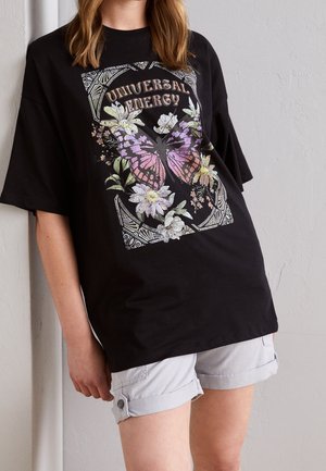 Personne portant un t-shirt noir oversize avec un papillon rose et des fleurs blanches en graphique et le texte "Universal Energy", accompagné d'un short clair.
