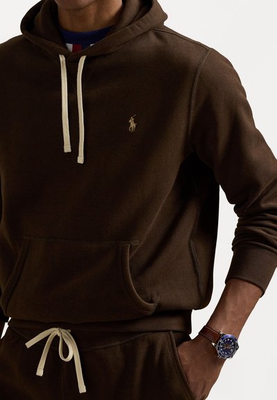 Rjava kapuca iz mehkega materiala s sprednjim žepom, opremljena s kremnim vezalcem in manjšim zlatim logotipom. Na zapestju je ura.