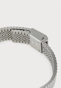 Bracelet de montre en métal avec un design tissé, finition en argent brillant et une fermeture à clip avec des détails gravés sur le matériel.