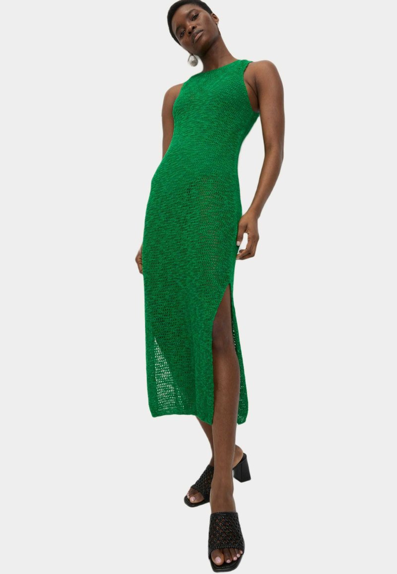 Grünes Strick-Maxikleid mit figurbetontem Design, seitlichem Schlitz und strukturierter Stoffqualität. Kombiniert mit schwarzen Blockabsatzschuhen mit offenen Zehen.