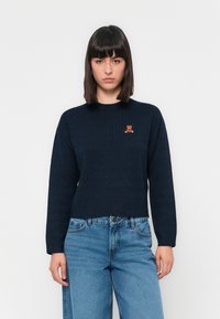 Granatowy sweter w warkoczowy splot z długimi rękawami i okrągłym dekoltem, z małym haftowanym logo misia na piersi. W połączeniu z niebieskimi dżinsami.
