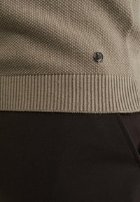 Béžový pletený svetr s žebrovaným lemem, s malým černým emblémem na přední straně. Tmavě hnědá látka viditelná dole.