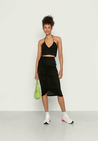 Top cropped preto com decote halter, combinado com uma saia midi preta drapeada. Usando sapatilhas brancas com detalhes em rosa e segurando uma bolsa verde.