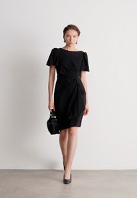 Melnas kleitas ar īsām pūkainām piedurknēm, asimetrisku drapējumu un pieguļošu siluetu. Aksesuāri ietver mazu melnu rokassomu un augstpapēžu kurpes.