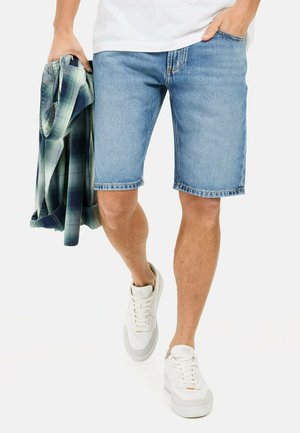 Lichtblauwe denim shorts met een rechte snit en een rafelige zoom. Draagt een witte t-shirt en witte sneakers, terwijl hij/zij een geruite overhemd vasthoudt.