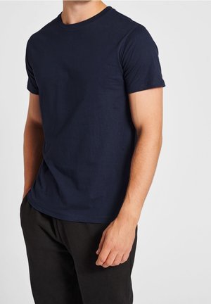 Homme portant un t-shirt bleu marine uni à manches courtes et un pantalon noir, debout, la main droite détendue le long du corps, sur un fond clair.
