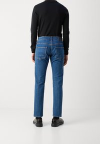 Raka, medelhöga blå denimbyxor med bakfickor, kombinerade med en svart långärmad tröja och svarta skor, visade från baksidan.