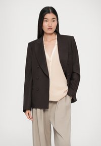 Blazer marrón de doble botonadura con solapas en pico, combinado con un cárdigan beige claro y pantalones beige holgados. Textura de tela suave.