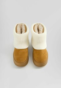 Bottes en daim marron clair avec un revers en maille côtelée crème. Doublure douce et moelleuse visible en haut. Bout rond et semelle en caoutchouc plate.