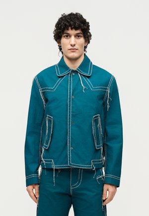 Νέος άνδρας με σγουρά μαλλιά φορώντας τζελ μπουφάν και ταιριαστό παντελόνι με λευκά εμφανή ραφές και χαλαρές κλωστές, στέκεται μπροστά σε απλό φόντο.