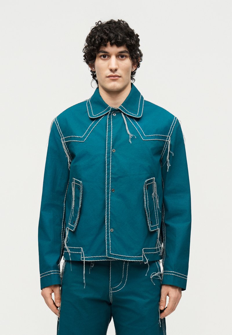 Νέος άνδρας με σγουρά μαλλιά φορώντας τζελ μπουφάν και ταιριαστό παντελόνι με λευκά εμφανή ραφές και χαλαρές κλωστές, στέκεται μπροστά σε απλό φόντο.