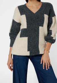 Pull en maille épaisse gris foncé et crème avec un col en V, des motifs géométriques contrastants, et des manches amples texturées.