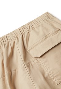 Béžové kalhoty s elastickým pasem a kapsou na flap vzadu, ukazující detailní švy a texturu látky.