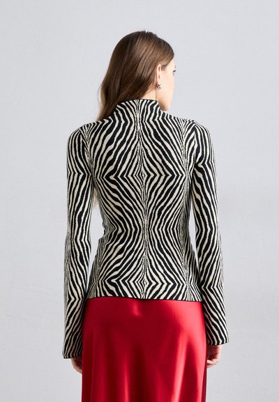 Ζαχαρωπή ζακέτα σε μαύρο και κρέμ, με ψηλό λαιμό, μακριά μανίκια και εφαρμοστή σιλουέτα, που συνδυάζεται με κόκκινη φούστα.