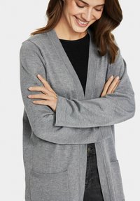 Cardigan grigio con maniche lunghe, apertura frontale, due tasche anteriori e polsini a costine, indossato sopra un top nero. Materiale in maglia morbido e texurizzato.