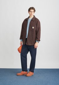 Model iført en oversized jakke med brune og orange mønstre, lys blå sweater, marineblå bukser og orange sko, mens hun holder en orange kasket.