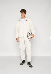 Hvit atletisk treningsoverallsdress med glidelåsjakke og bukser, begge med subtile aksenter. Modellen holder en svart-hvitt fotball.