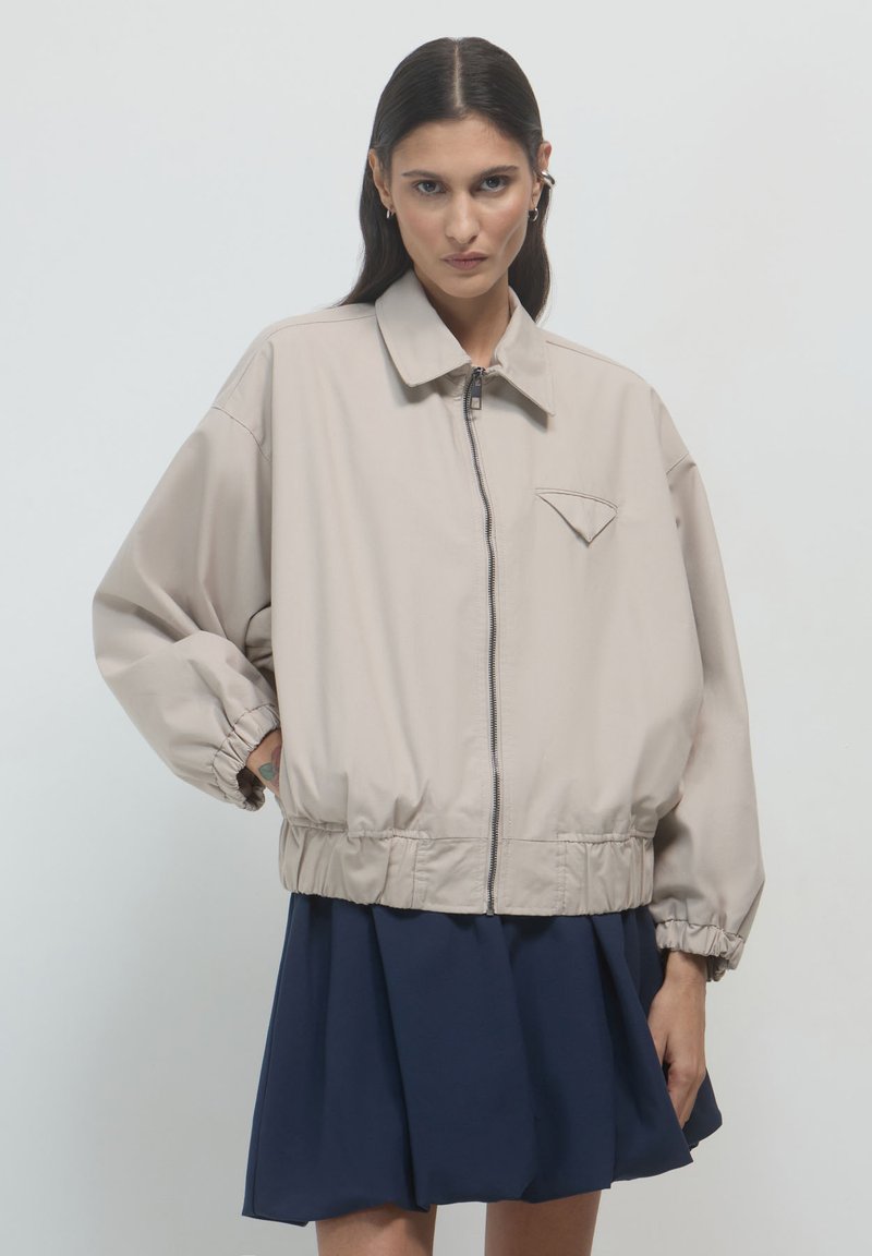 Hellbeige Jacke mit Reißverschluss, spitzem Kragen und elastischem Bund. Mit gekrempelten Ärmeln und einer kleinen Brusttasche.