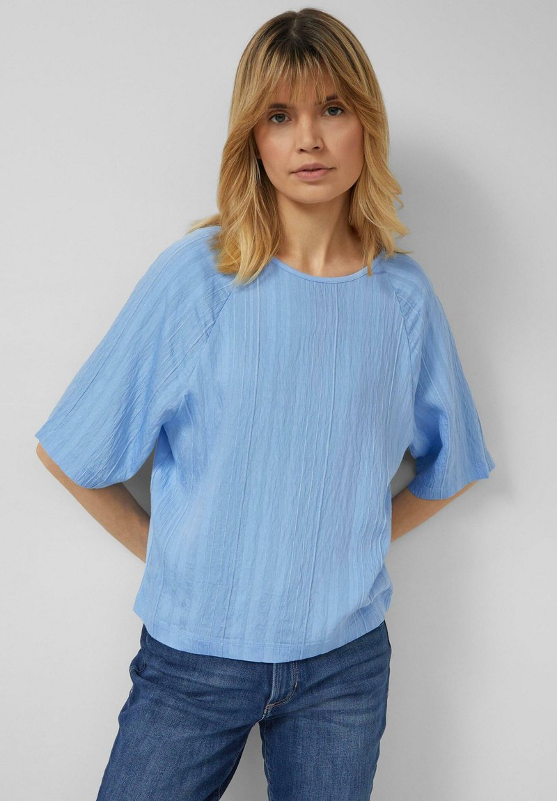 Blond žena v lehkém modrém texturovaném volném krátkém rukávu a modrých džínách stojící před jednobarevným světle šedým pozadím.