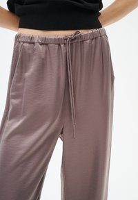 Lehké, mauve kalhoty s elastickým pasem a šňůrkou. Mají boční kapsy a hladkou, měkkou texturu.