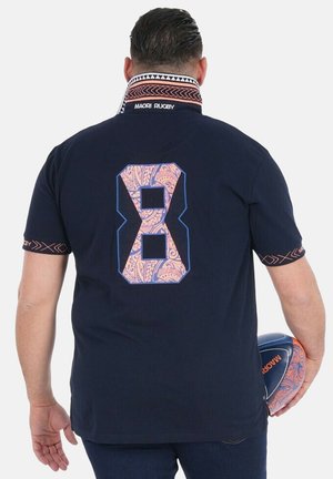 Homme portant un maillot de rugby maori bleu marine avec le numéro 8 en motif polynésien, tenant un ballon de rugby assorti derrière son dos.