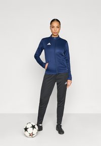 Ναυτικό αθλητικό μπουφάν με φερμουάρ και όρθιο γιακά, συνδυασμένο με μαύρο αθλητικό παντελόνι. Φέρει το λογότυπο της Adidas· το μοντέλο στέκεται δίπλα σε μαύρη και λευκή ποδοσφαιρική μπάλα.