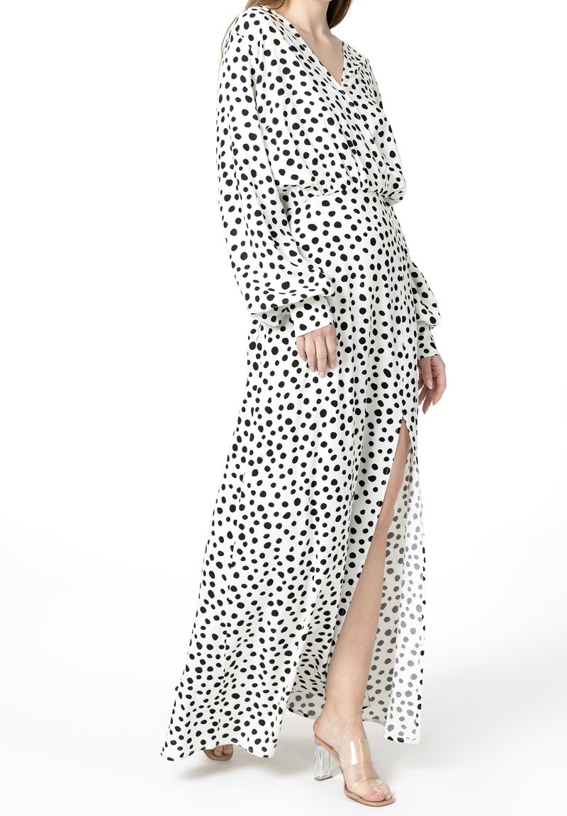 Femme portant une longue robe blanche à pois noirs, manches longues, col en V et fente haute sur la cuisse, associée à des sandales à talons hauts transparentes.