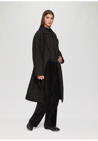 Oversized zwarte wollen jas met een gestructureerde stof, brede revers en twee knopen. Gecombineerd met zwarte brede broek en gestikte zwarte schoenen.