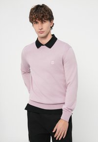 Mladi moški s kodrastimi lasmi, oblečen v svetlo vijoličen pulover čez črno srajco z ovratnikom in črne hlače, stoji pred belim ozadjem.