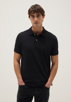 Giovane uomo con capelli castani corti che indossa una polo nera a maniche corte e pantaloni neri, in piedi con le mani nelle tasche.