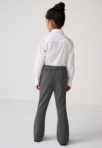 Mädchen mit dunkel gebundenem Haar, das ein weißes Langarmshirt und graue Hose mit elastischem Bund trägt, steht mit dem Rücken zu einer schlichten Wand.