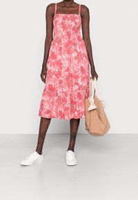 Robe midi rose à motif floral avec un corsage froncé, des bretelles fines et un bas évasé. Associée à des baskets blanches et un sac beige.