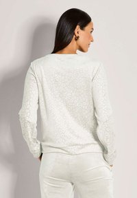 Frau mit langen dunklen Haaren, die ein weißes, strukturiertes Langarmoberteil und passende Hose trägt, seitlich blickend mit Händen in den Taschen.