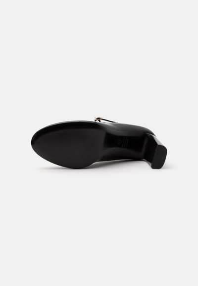 Chaussure à talon en cuir noir vue de la semelle, avec un talon carré et une petite boucle métallique sur la partie supérieure.