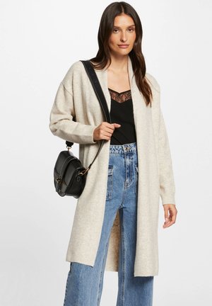 Cardigan di maglia color crema con apertura frontale, tessuto testurizzato, maniche lunghe e spacchi laterali, abbinato a un top nero e jeans a vita alta.