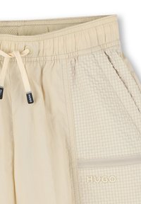 Beige drawstring shorts with a smooth texture. Features a grid-patterned panel and a zippered pocket. "HUGO" logo in light embroidery.