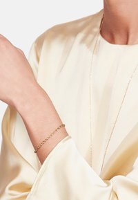 Kullaketju, joka on kierretty kultaiseksi, on käytössä ranteessa ja sen kera on vaalea satiininen pitkähiha blouse. Hienovarainen kiilto korostaa kankaan rakennetta.