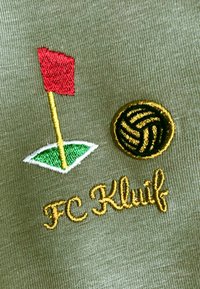 Geborduurde patch op olijfgroen stof met een rode vlag op een groene golftee en een zwart-gouden volleyball, gelabeld "FC Kluif."
