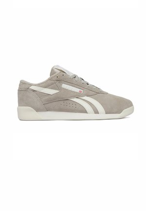 Zapatilla deportiva de ante gris claro con rayas, cordones y suela blancas, vista lateral con detalle de una pequeña bandera cerca de los cordones.