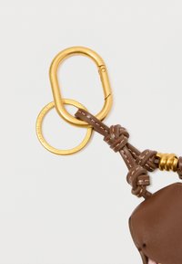 Pruun nahast võtmehoidja, millel on kuldne riistvara. Omab karabiini klambrit ja ümmargust rõngast, mis on kindlalt seotud kudumisega nahavööde abil.